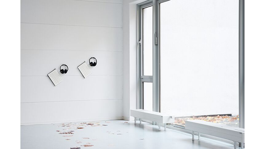 In an exhibition space tactile paintings of crunchy leaves on the floor mirror some leaves outside of the window. Hung on the wall are two headphones and accompanying access copies of a sound artwork.