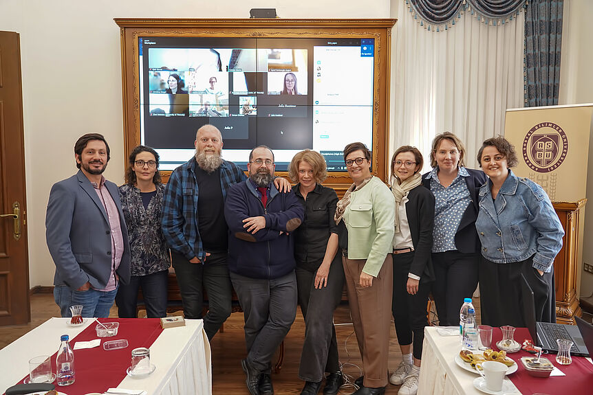 Nine persons standing in front of a large screen displaying a Zoom room with thirteen camera windows