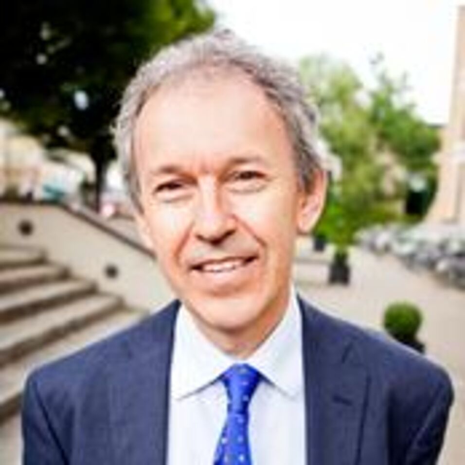 the face of a man wearing a black suit and a blue tie in an outdoor setting, trees and a staircase in the background