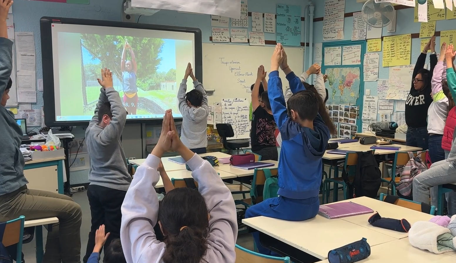 Students in a classroom. They are facing a smartboard with raised hands to stretch their bodies